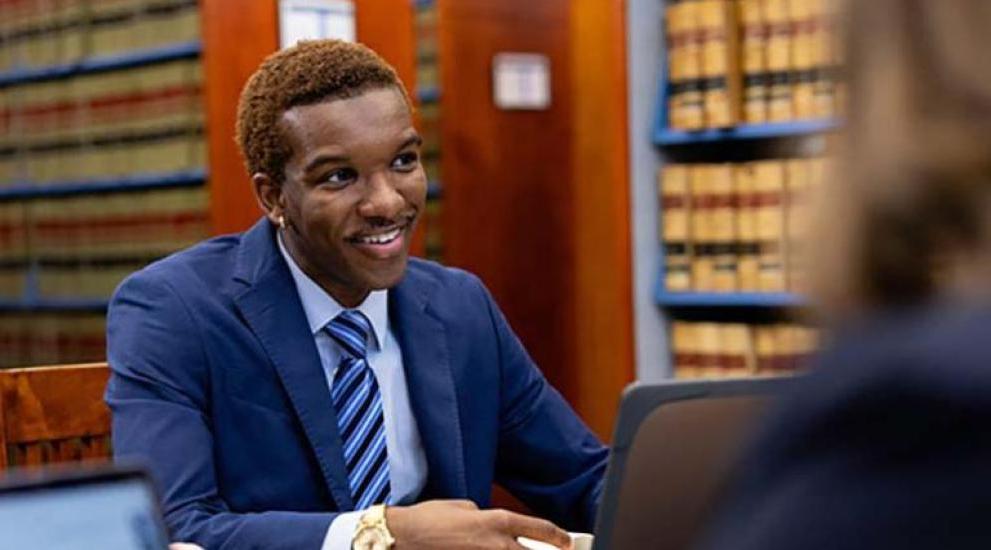 Law student dressed in business suit sitting in law library talking to another student.